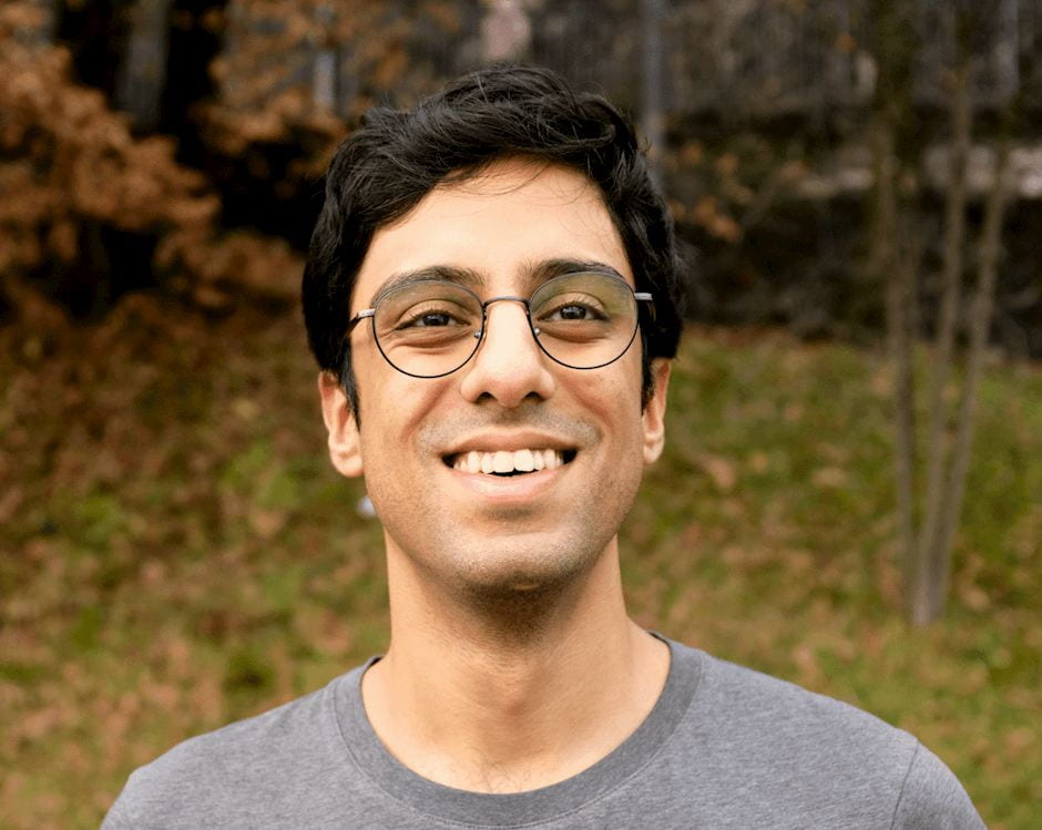Picture of Manav smiling and standing in front of a leaf-covered hill